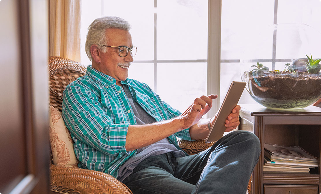 Older man smiling at a tablet in a wicker chair.